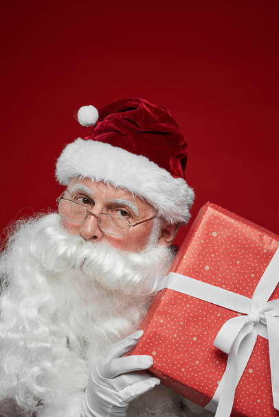Portrait du Père Noël avec barbe blanche et chapeau posant à la caméra avec boîte cadeau rouge
 - Photo, image
