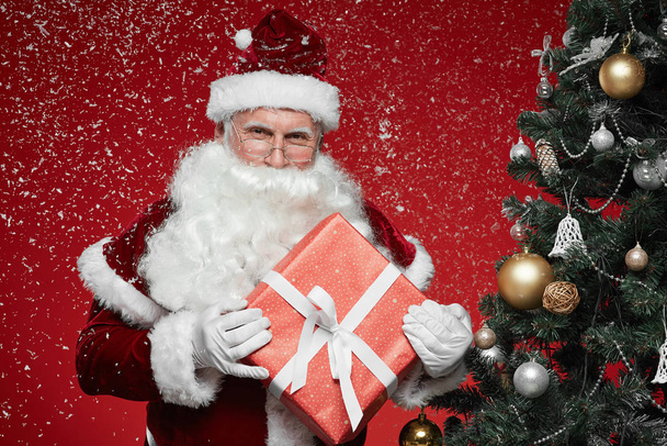 Portrait of Santa Claus in red costume holding Christmas present while standing near the Christmas tree during snowfall - Photo, image