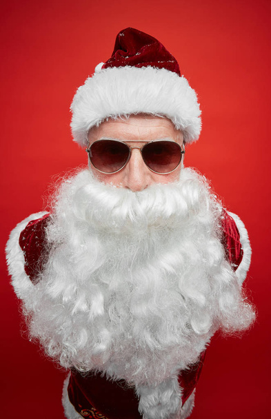 Porträt eines modernen Weihnachtsmannes mit weißem Bart, der eine stylische Sonnenbrille trägt und vor rotem Hintergrund in die Kamera blickt - Foto, Bild