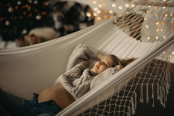 Pregnant woman rests in hammock against background of Christmas decorations - 写真・画像
