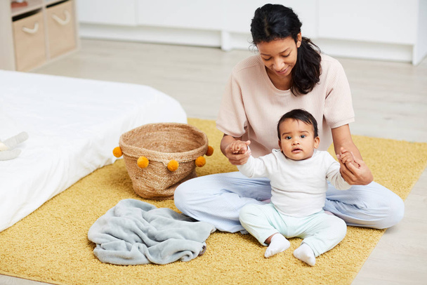 Nuori onnellinen äiti istuu lattialla yhdessä vauvan kanssa kädestä ja auttaa istumaan.
 - Valokuva, kuva