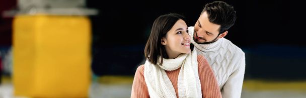panoramiczne ujęcie młodej uśmiechniętej pary w swetrach ściskającej się na lodowisku  - Zdjęcie, obraz