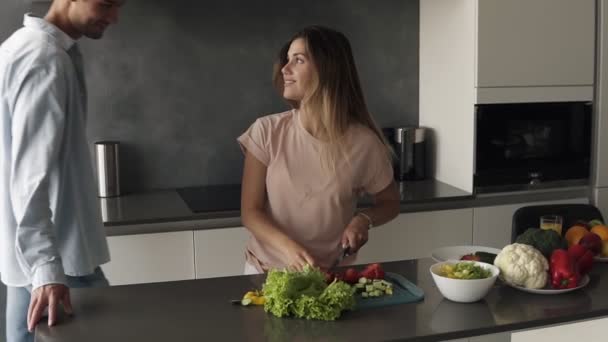 A young man is kissing his girlfriend when shes making a fresh salad for them. He came to hug her and appreciate. Long haired woman kisses him back. Modern kitchen in grey colours - Filmati, video