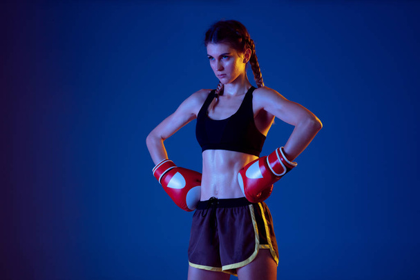 Fit caucasian woman in sportswear boxing on blue studio background in neon light - Фото, изображение