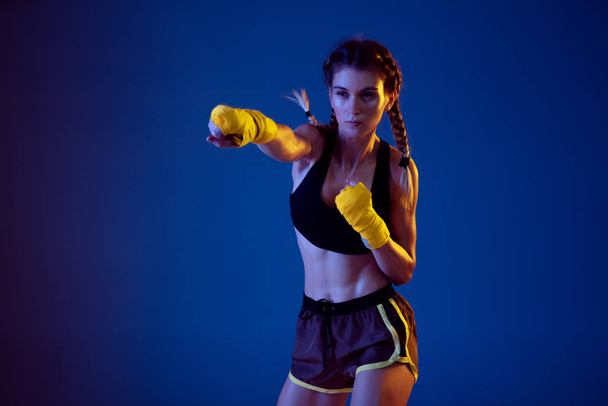 Fit caucasian woman in sportswear boxing on blue studio background in neon light - Foto, Imagem