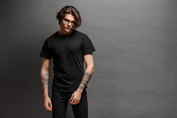 Hipster handsome male model with glasses wearing black blank t-shirt and black jeans with space for your logo or design in casual urban style - Fotó, kép