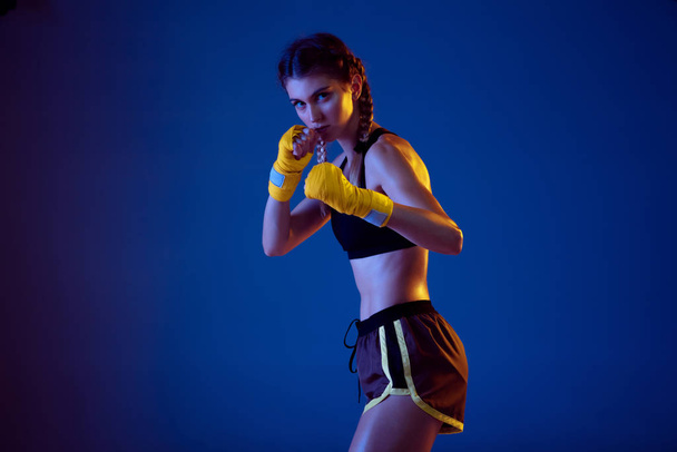 Fit caucasian woman in sportswear boxing on blue studio background in neon light - Фото, изображение