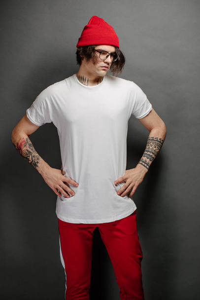 Hipster handsome male model with glasses wearing white blank t-shirt and red pants and a red hat with space for your logo or design over gray background. - Fotografie, Obrázek