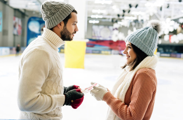 knappe man geven wenskaart op Valentijnsdag aan vrolijke vrouw op schaatsbaan - Foto, afbeelding