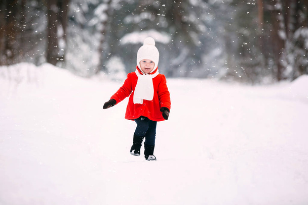 hezká holčička v červeném kabátě v zimním lese. Holčička se v zimě baví. veselé holčička v rukavicích a bílý klobouk běží na sněhobílé - Fotografie, Obrázek