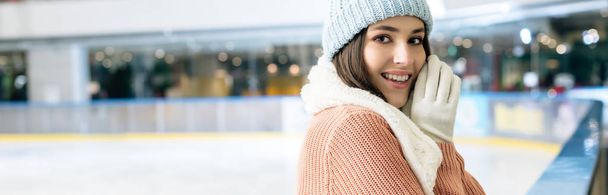 plano panorámico de chica atractiva alegre en suéter, bufanda, guantes y sombrero de pie en la pista de patinaje
 - Foto, Imagen