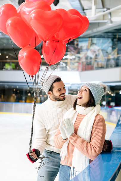 ενθουσιασμένοι ζευγάρι σε χειμερινό ντύσιμο με κόκκινο μπαλόνια σχήμα καρδιάς ξοδεύουν χρόνο για πατινάζ παγοδρόμιο την ημέρα του Αγίου Βαλεντίνου - Φωτογραφία, εικόνα