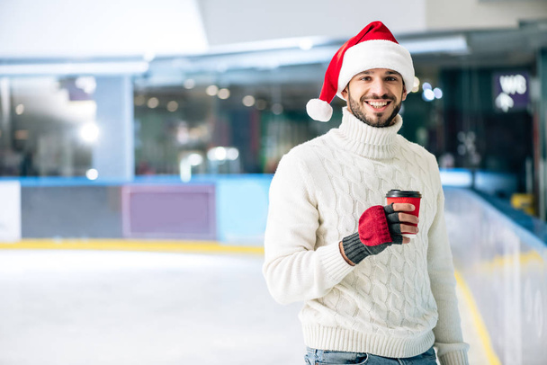 Χαρούμενος άντρας με πουλόβερ και καπέλο Σάντα που κρατάει καφέ για να πάει για πατινάζ - Φωτογραφία, εικόνα