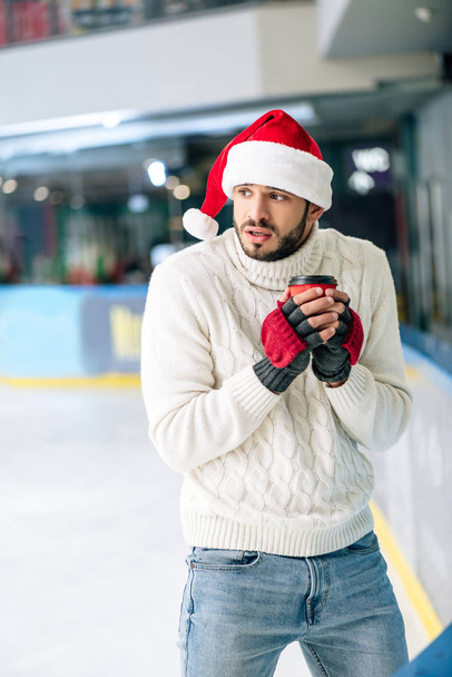 Süveter ve Noel Baba şapkalı soğuk adam paten pistine gitmek için elinde kahve tutuyor. - Fotoğraf, Görsel