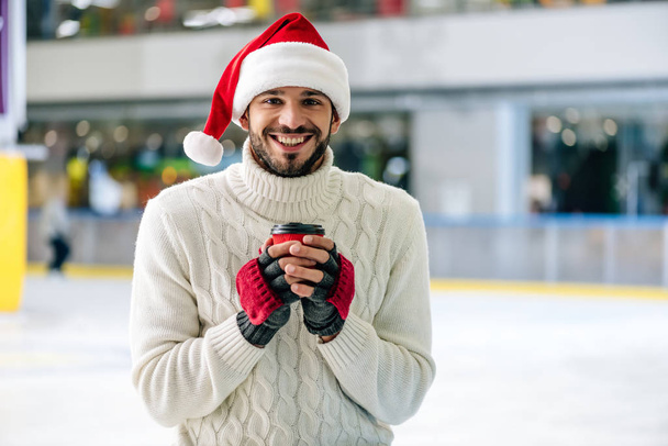 handsome cheerful man in santa hat holding coffee to go on skating rink - Foto, afbeelding