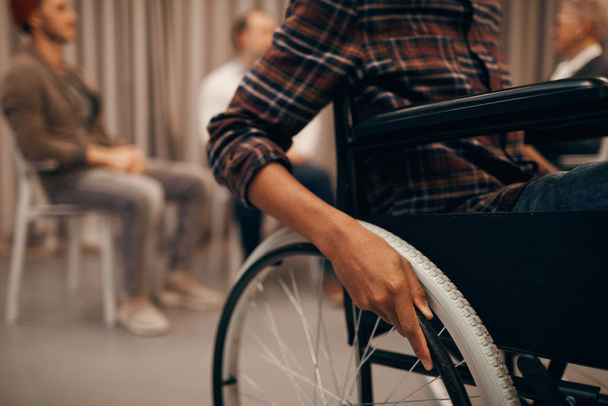 Arka planda bir grup insanla birlikte tekerlekli sandalyede oturan genç adamın yakın çekimi. - Fotoğraf, Görsel