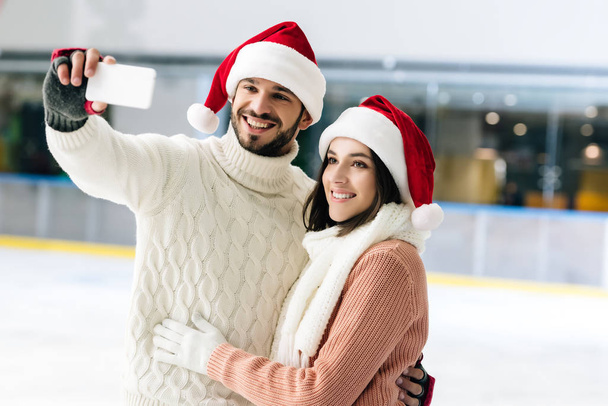 smiling couple in santa hats taking selfie on smartphone on skating rink at christmastime - Foto, Bild