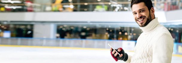 plano panorámico de hombre sonriente guapo usando teléfono inteligente en pista de patinaje
 - Foto, imagen