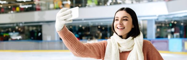 panoramisch schot van aantrekkelijke vrolijke meisje het nemen van selfie op smartphone op schaatsbaan - Foto, afbeelding