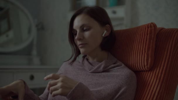 Brunette female taking wireless earphones to her ears and enjoying the music. Close up of woman in dress sitting in cosy orange armchair in bedroom.  - Imágenes, Vídeo