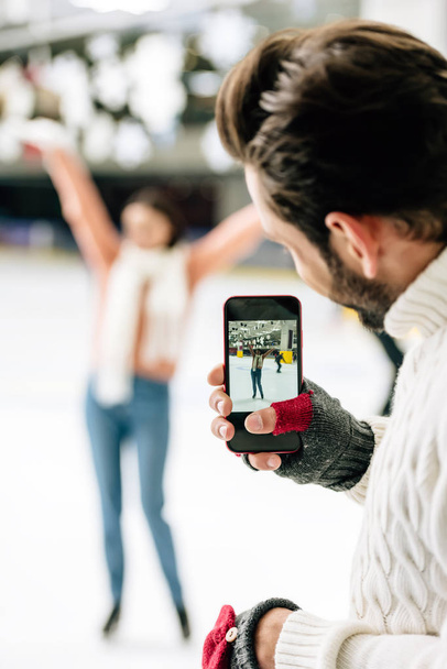 επιλεκτική εστίαση του όμορφος άντρας λήψη φωτογραφιών του ενθουσιασμένος γυναίκα στο smartphone για παγοδρόμιο - Φωτογραφία, εικόνα