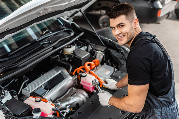 handsome mechanic inspecting car engine compartment and smiling at camera - Photo, Image