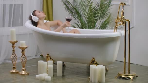Close up of young woman lying in a bathtub listening to music with big headphones on her head and drink red wine from a glass. Concept of relaxation and freedom - Footage, Video