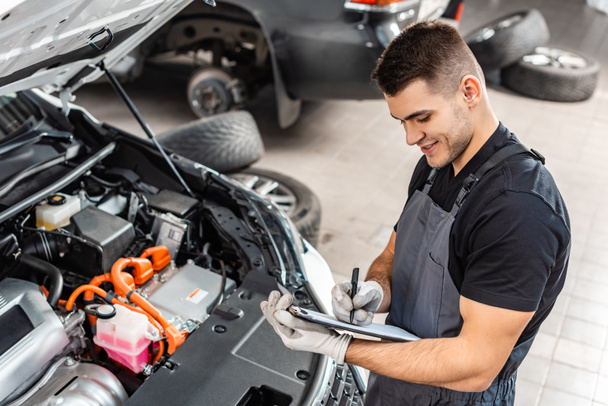smiling mechanic writing on clipboard while inspecting car engine compartment - Photo, Image