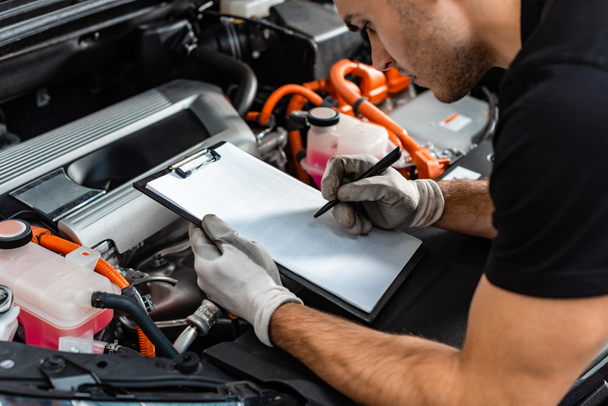 bijgesneden weergave van monteur schrijven op klembord tijdens de inspectie van de auto motorruimte - Foto, afbeelding