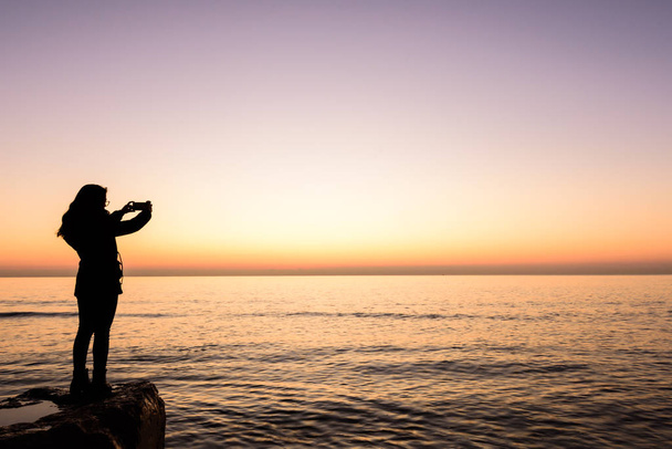 Девушка фотографирует с мобильного телефона восход солнца на пляже Оропеса, Валенсия, Испания
 - Фото, изображение