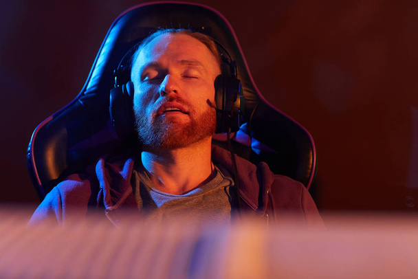 Młody brodaty mężczyzna w słuchawkach siedzi w fotelu do gier i śpi zmęczony po grze - Zdjęcie, obraz