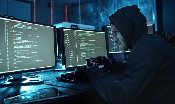 Jovem hacker de computador com capuz olhando para monitor de computador e quebrando o software no escritório escuro
  - Foto, Imagem