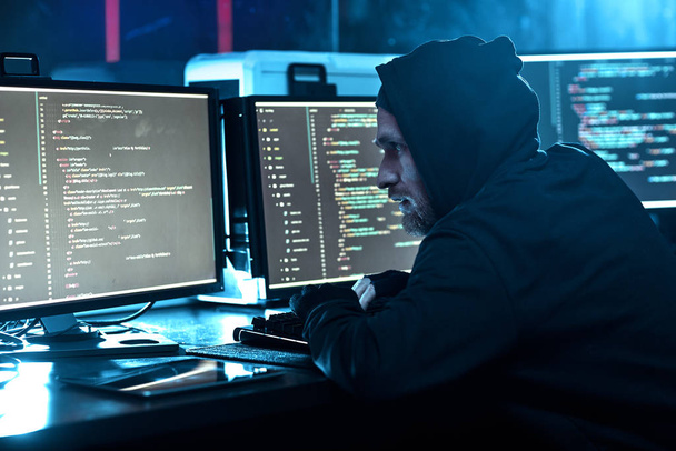 Mladý počítačový hacker v Hoody při pohledu na monitor počítače a lámání software v tmavé kanceláři  - Fotografie, Obrázek