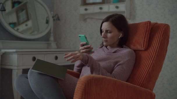 Молодая женщина читает бумажную книгу и фотографирует страницу на смартфоне для проверки в приложении. Брюнетка с камерой и книга сидя в уютном оранжевом кресле в спальне
 - Кадры, видео