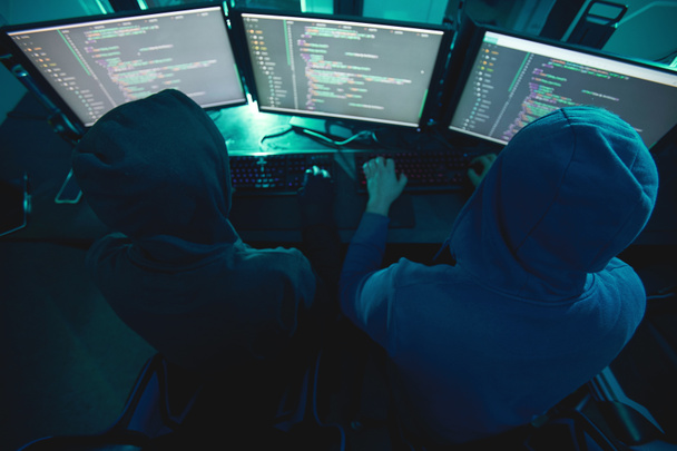コンピュータ監視の前に座って、システムを破壊し、チームで作業するコンピュータのハッカーの背面ビュー - 写真・画像