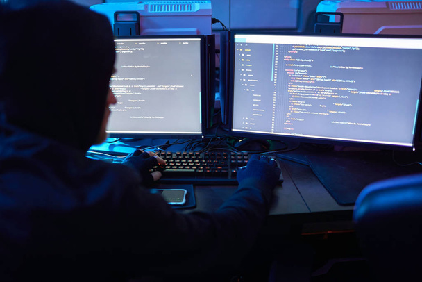 Rückansicht eines Computerhackers mit Kapuzenpulli, der am Tisch vor Computermonitoren sitzt und im dunklen Büro Computercodes bricht - Foto, Bild