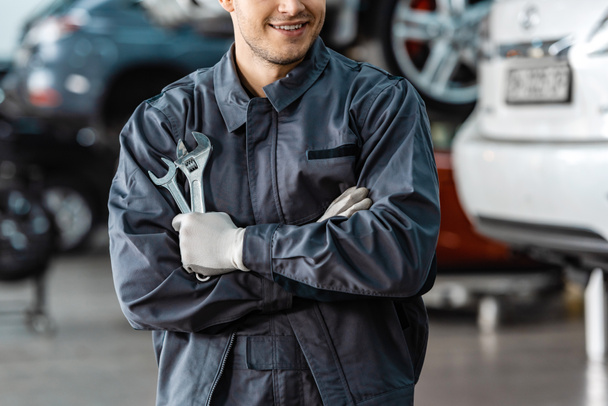 oříznutý pohled na usmívajícího se mechanika stojícího se zkříženýma rukama a držícího klíče - Fotografie, Obrázek