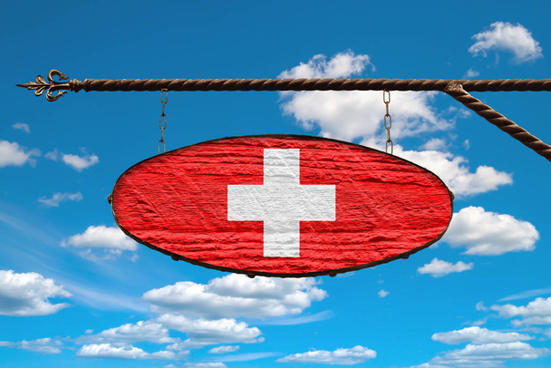 Switzerland flag on signboard. Oval signboard colors flag Switzerland hangs on a metal forged structure. Template on a background of blue sky with clouds.  - Zdjęcie, obraz