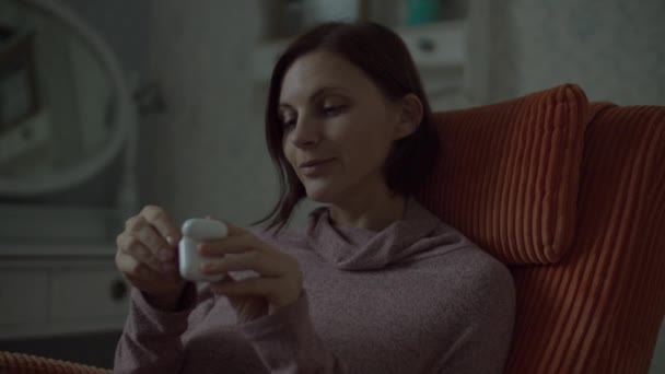 Brunette female taking wireless earphones to her ears and enjoying the music. Close up of woman in dress sitting in cosy orange armchair in bedroom.  - Záběry, video