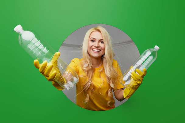 Jonge vrouwelijke vrijwilliger in gele handschoenen met recycling bak met plastic afval kijkt door het ronde gat in een groene achtergrond. Milieubeschermingsconcept - Foto, afbeelding