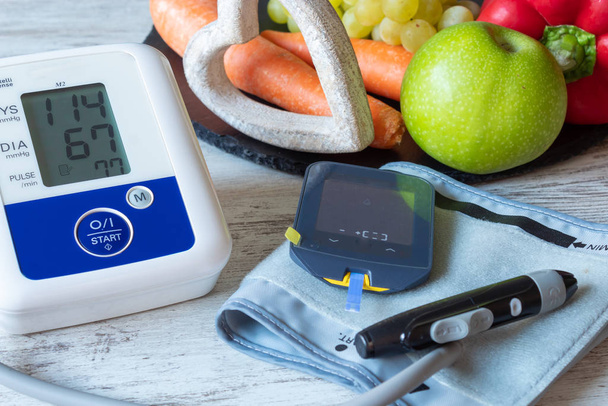 Приборы для измерения артериального давления и глюкозы крови на столе с фруктами и овощами
 - Фото, изображение