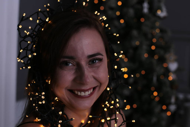 Beautiful girl with short dark hair with a garland near a Christmas tree decorated - Фото, изображение