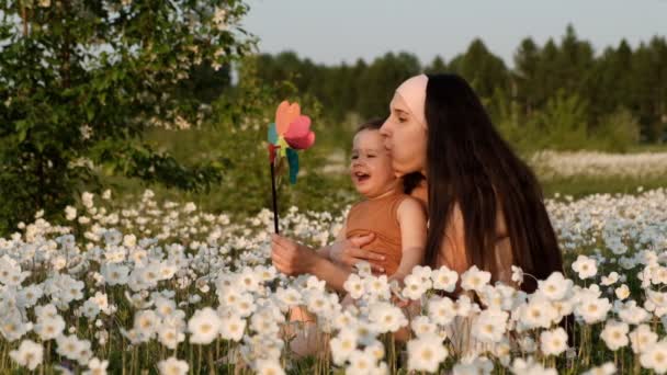 Медленное движение молодой привлекательной матери с сыном, играющим на ветряной мельнице, сидящей на цветочном поле на закате
. - Кадры, видео