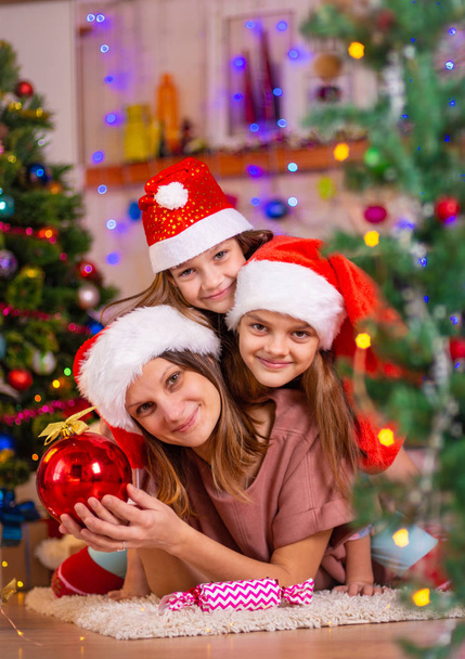 Mom and two daughters lie on the floor of a Christmas tree in the New Year's interior - Фото, изображение