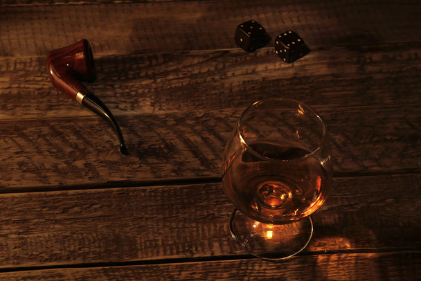 Glass with cognac or whiskey and smoking pipe on a wooden table. Dark and Moody style. - Foto, imagen