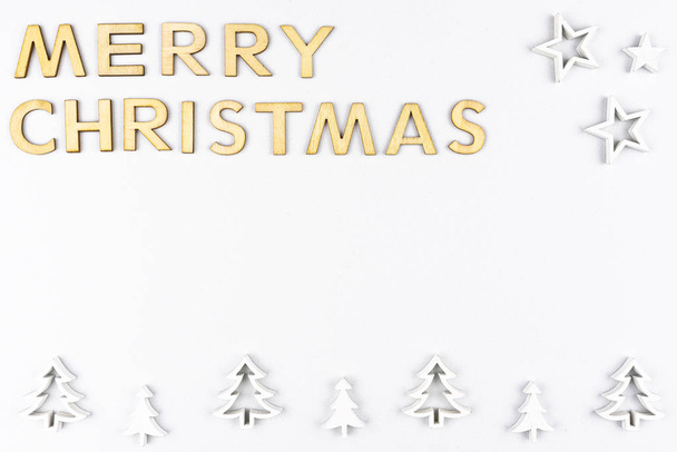 De inscriptie Vrolijk kerstfeest gemaakt van houten letters, plat van boven, geïsoleerd op een witte, ruwe achtergrond. Witte houten kerstbomen gerangschikt in een rij op de bodem. - Foto, afbeelding
