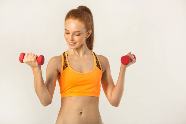 beautiful young woman with red hair with dumbbells in hands on a white background - Фото, изображение