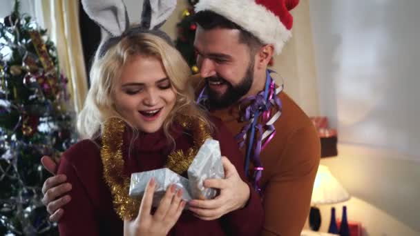 Young Caucasian blond woman in bunny ears headband opening gift box and hugging her adorable husband. Bearded man in Christmas hat making surprise present for wife on New Years eve. Holidays. - Séquence, vidéo
