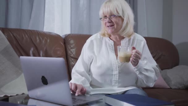 Positive Caucasian senior woman in eyeglasses sitting at couch with coffee and surfing internet. Blond mature retiree using laptop at home. Leisure activity, pastime. - Кадры, видео