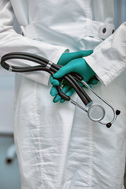Arzt hält Stethoskop hinter dem Rücken, hüfthohes Porträt auf Klinikhintergrund - Foto, Bild